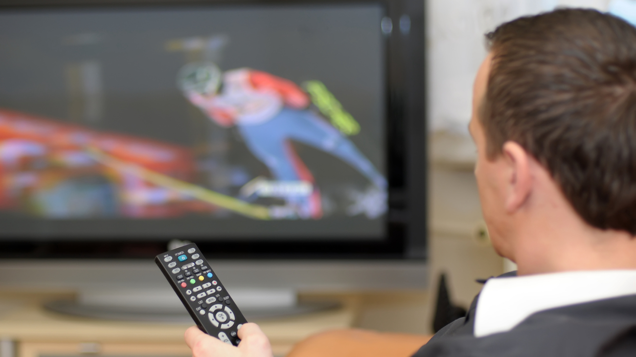 A man holding a remote control while watching a ski jumping on TV.